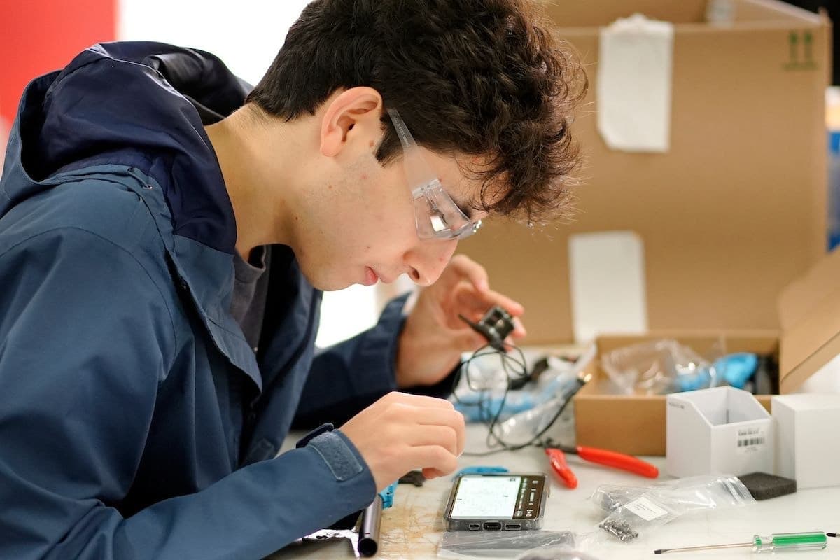 A participant wearing safety glasses examines a small wired component beside tools and drone-kit parts.